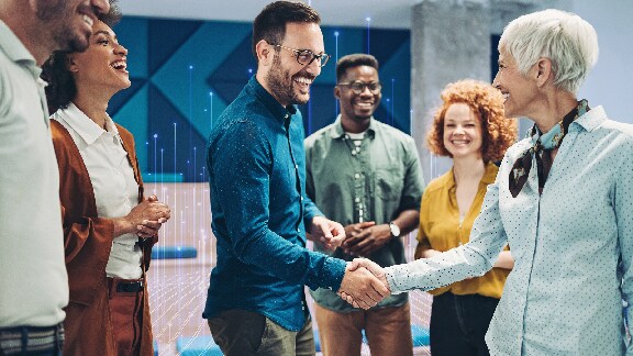 A man and a woman shaking hands, while their colleagues smile and watch around them